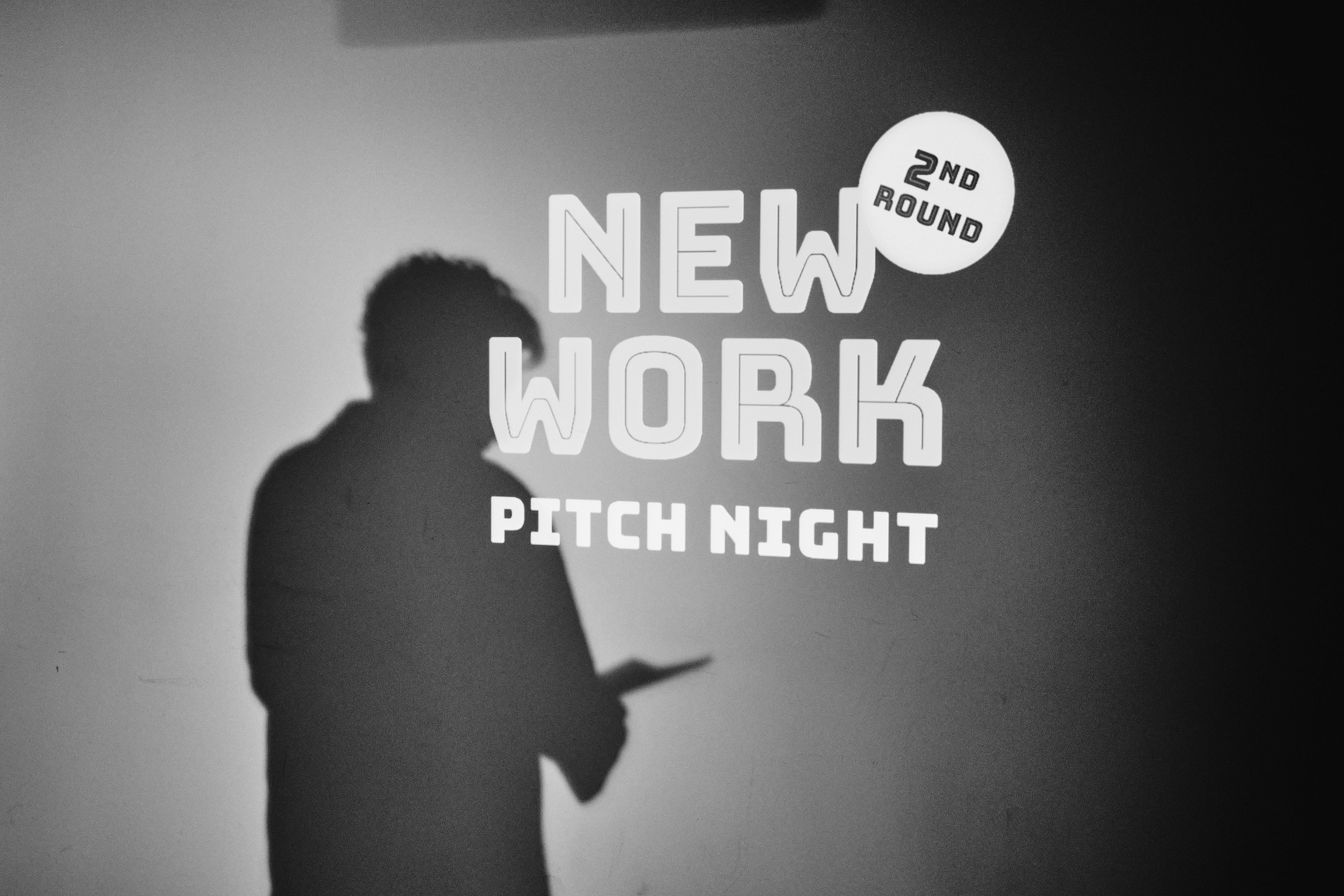 Der Schatten einer Person, die Papiere in der Hand hält, erscheint an einer Wand mit der Aufschrift "New Work Pitch Night 2nd Round".